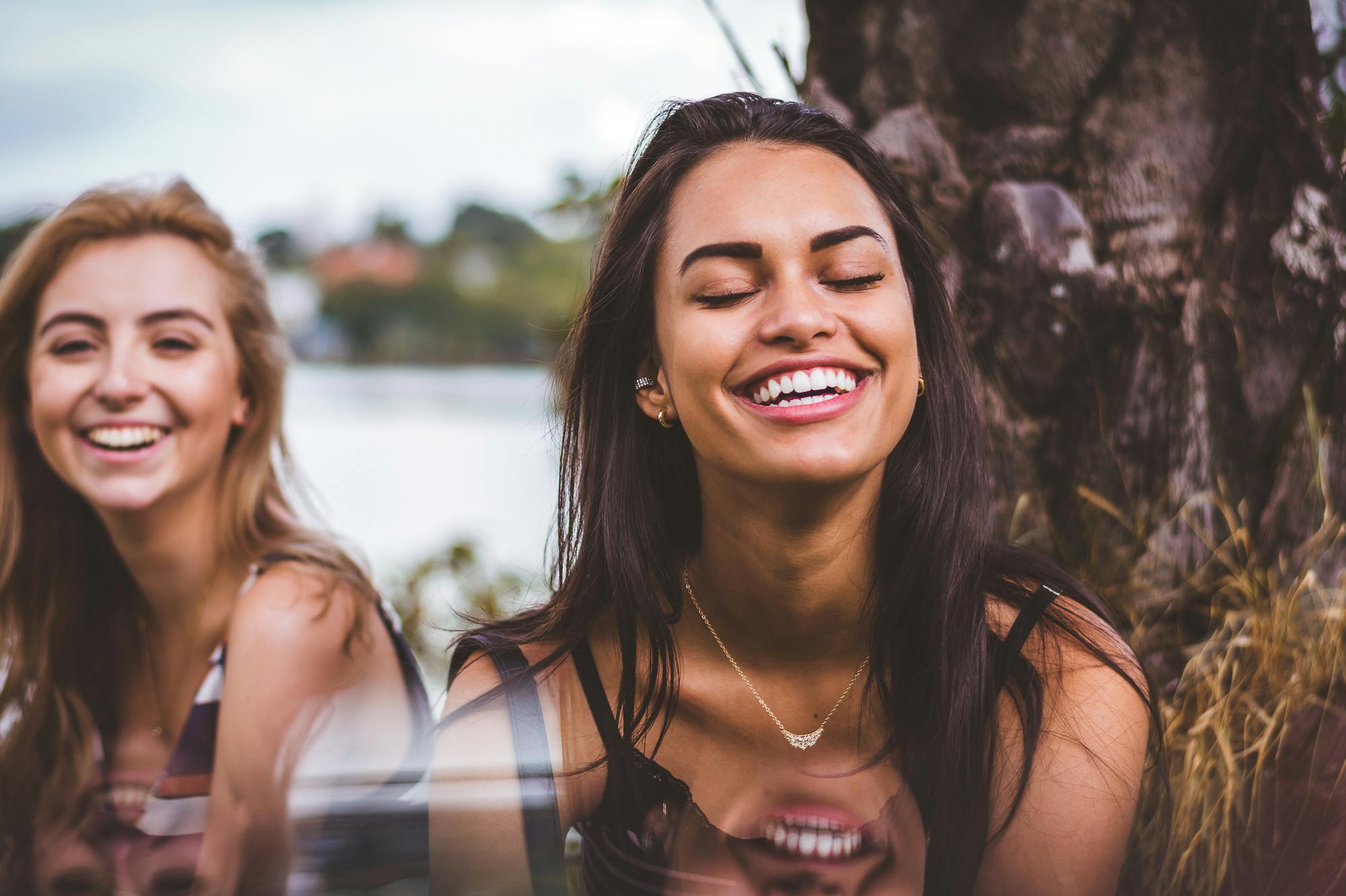 Services Two women happily laughing together outdoors by a serene lakeside.